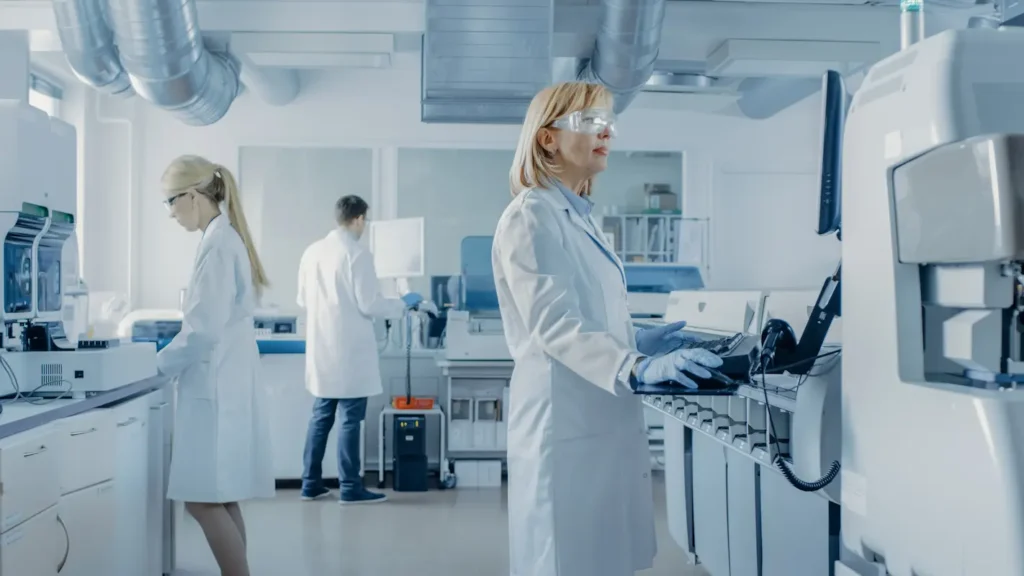 Three scientists work in a modern laboratory. A woman wearing safety goggles operates equipment in the foreground, while another woman and a man work in the background.