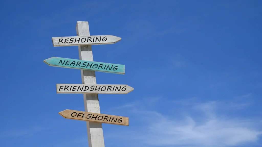 A signpost against a blue sky displays four directional arrows labeled: Reshoring, Nearshoring, Friendshoring, and Offshoring.