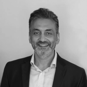 Smiling man in a suit with a striped shirt, grey beard, and short hair against a plain background.