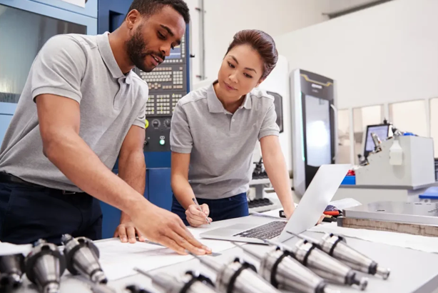 Two engineers collaborate in a modern factory, discussing blueprints with machinery in the background.