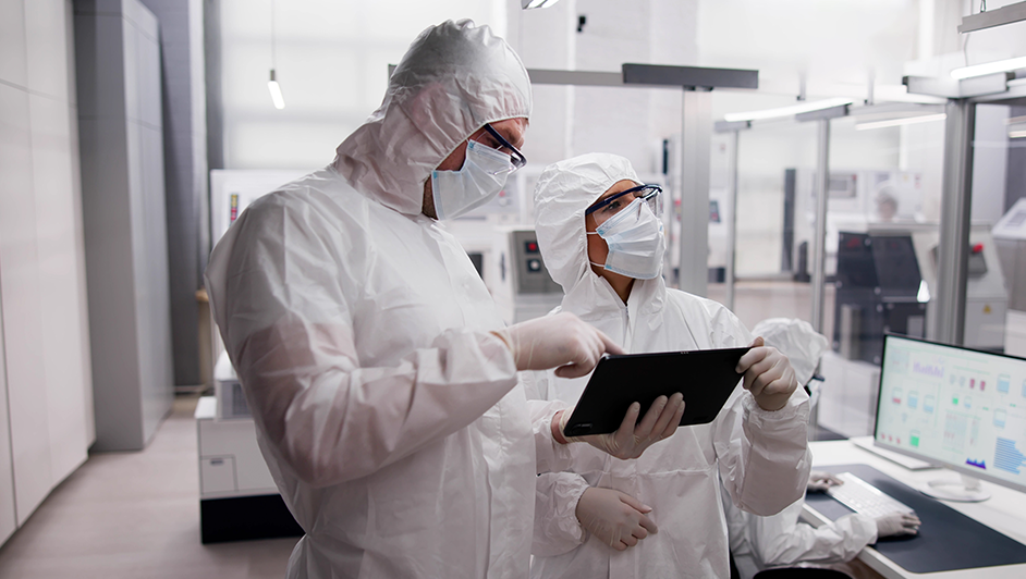 Scientists in cleanroom suits analyze data on a tablet in a laboratory.