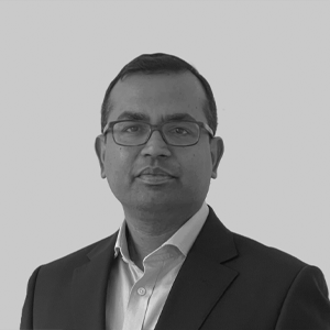 Portrait of a man in a suit and glasses, looking at the camera, set against a gray background.
